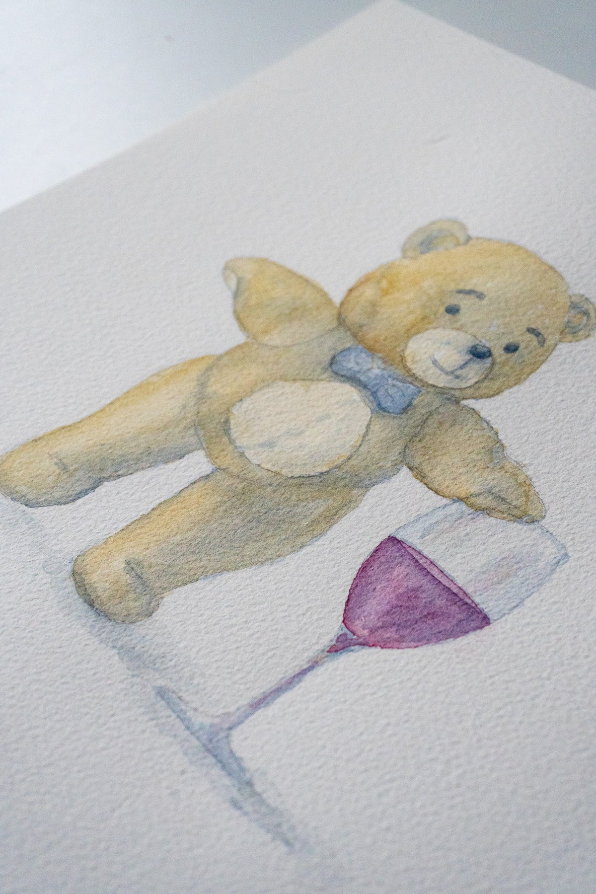 A watercolor-style illustration of a teddy bear lying beside a wine glass on textured white paper.